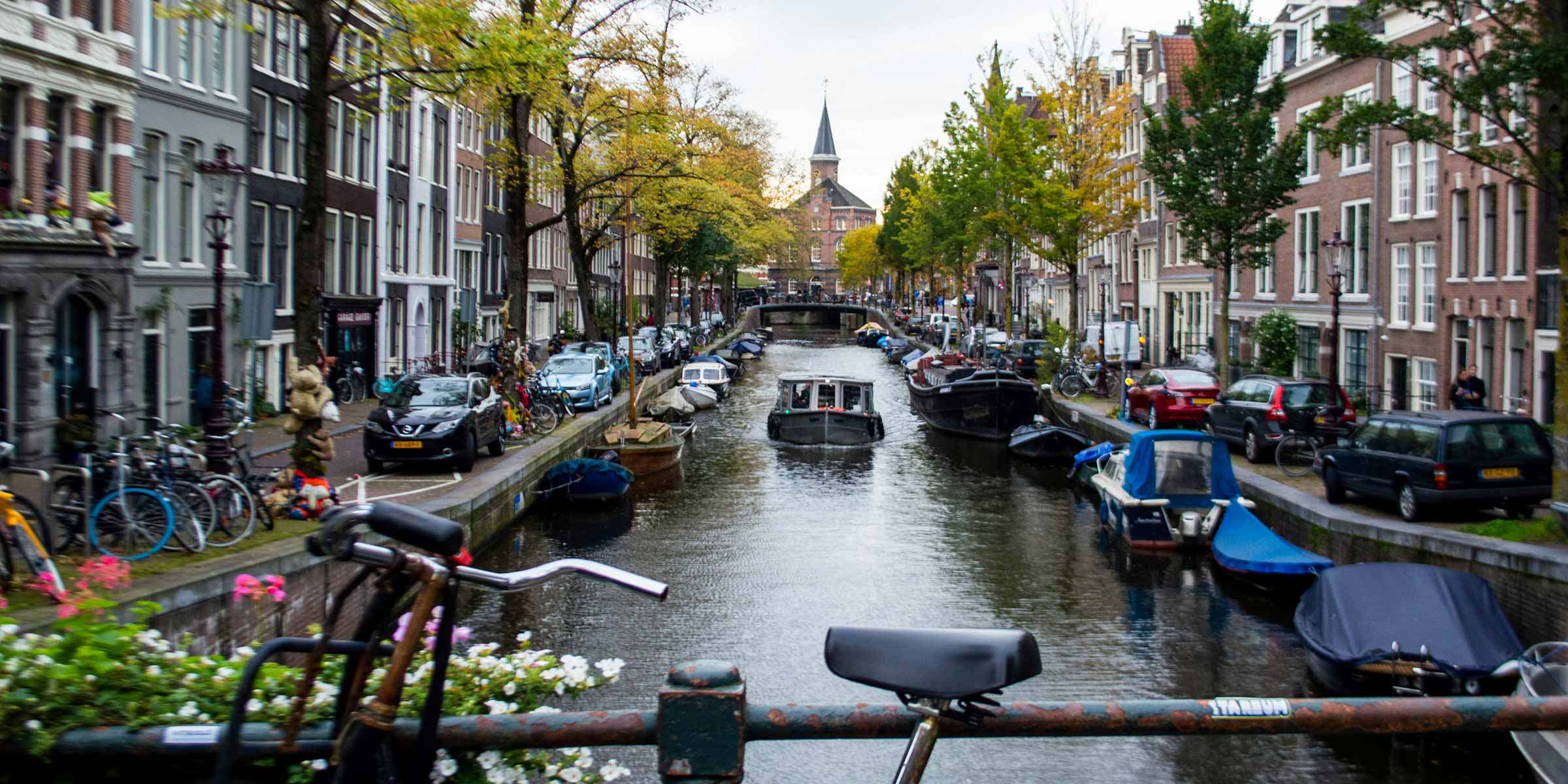 View from a bridge in Amsterdam of the waterway lined by boats with cars and bikes parked on either side.